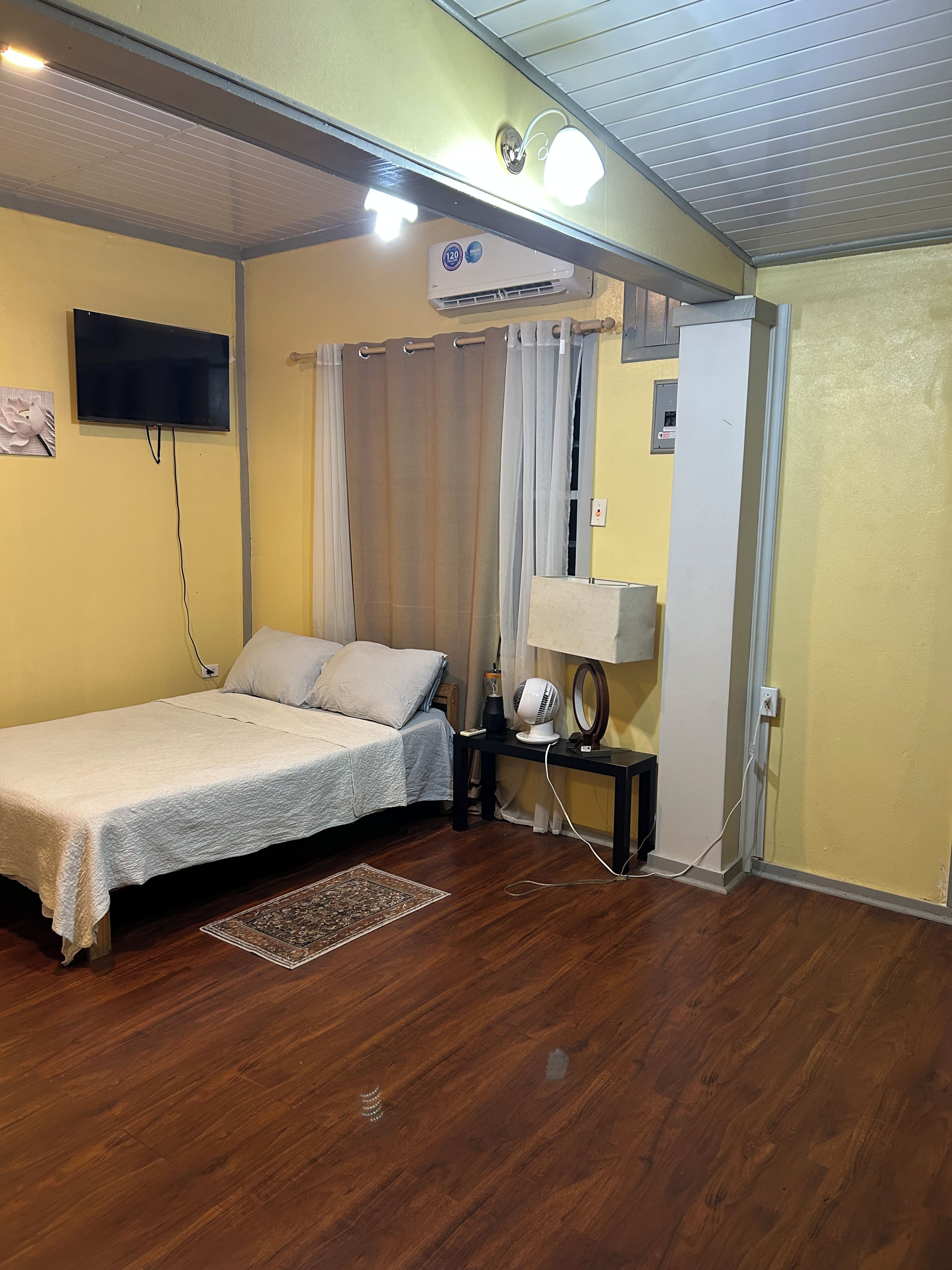 A room with a bed, a bedside table with a lamp, a wall-mounted TV, and an air conditioning unit.