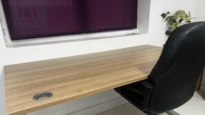 Un bureau en bois, une chaise de bureau noire, une fenêtre avec des stores violets et une petite plante posée sur le rebord de la fenêtre.