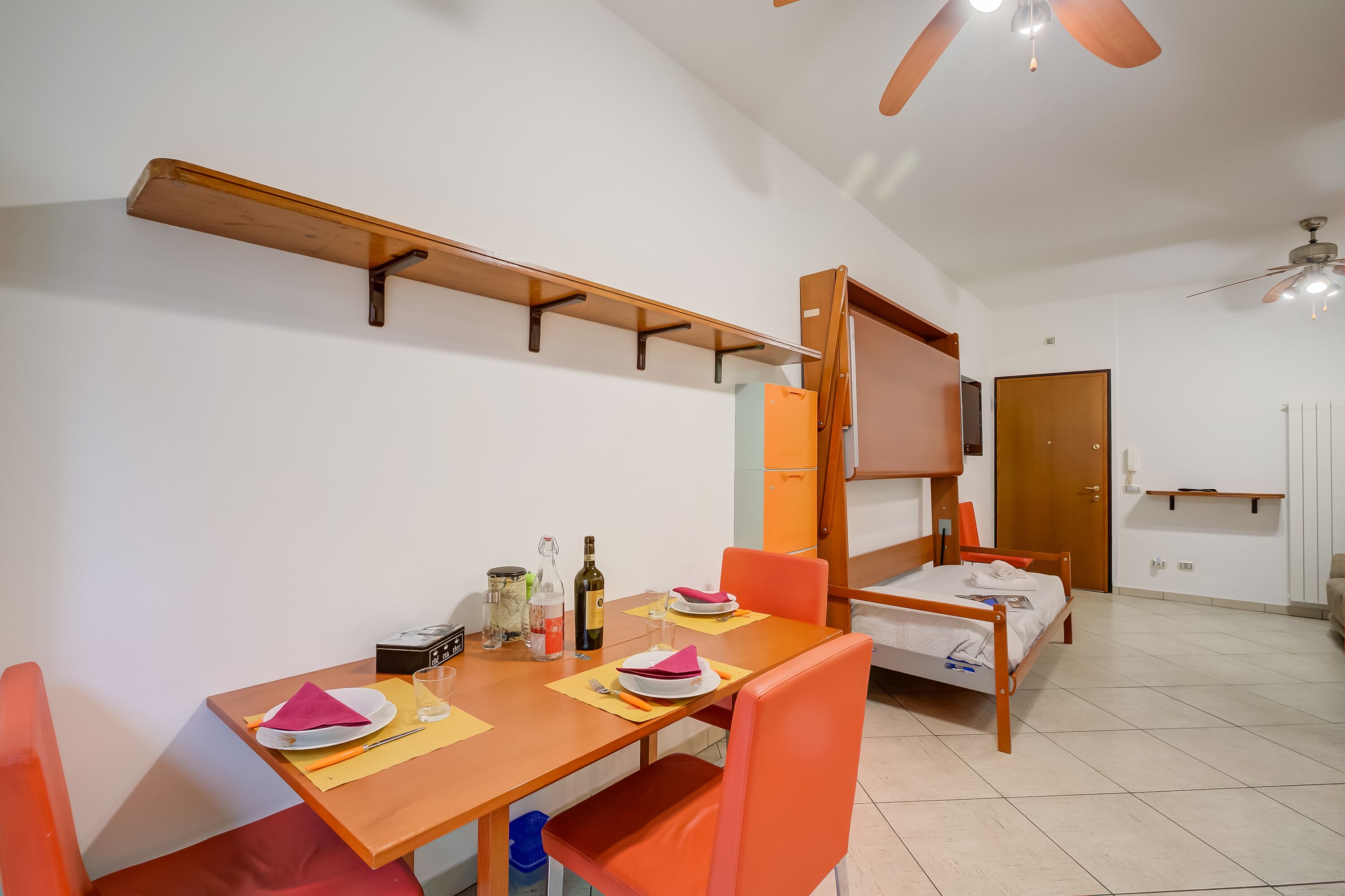 A compact room with a dining table set for two, a bed, a wardrobe, and a ceiling fan.