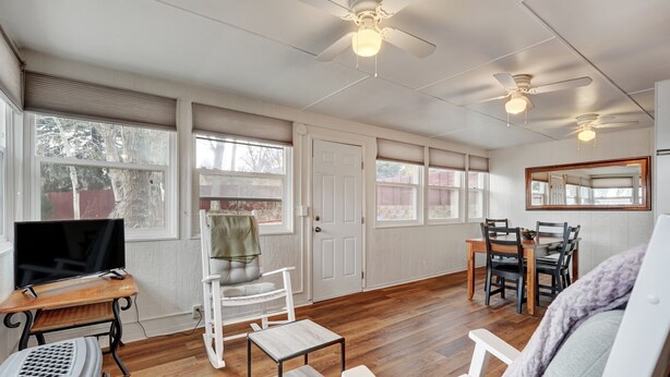 A spacious living room with a wooden floor, a TV on a wooden stand, a white armchair, a dining table with chairs, and a ceiling fan.