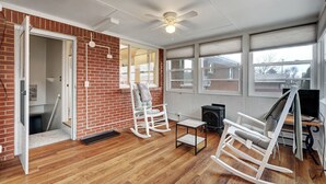 A room with wooden flooring, a brick wall, a ceiling fan, a rocking chair, a small table, and a fireplace.