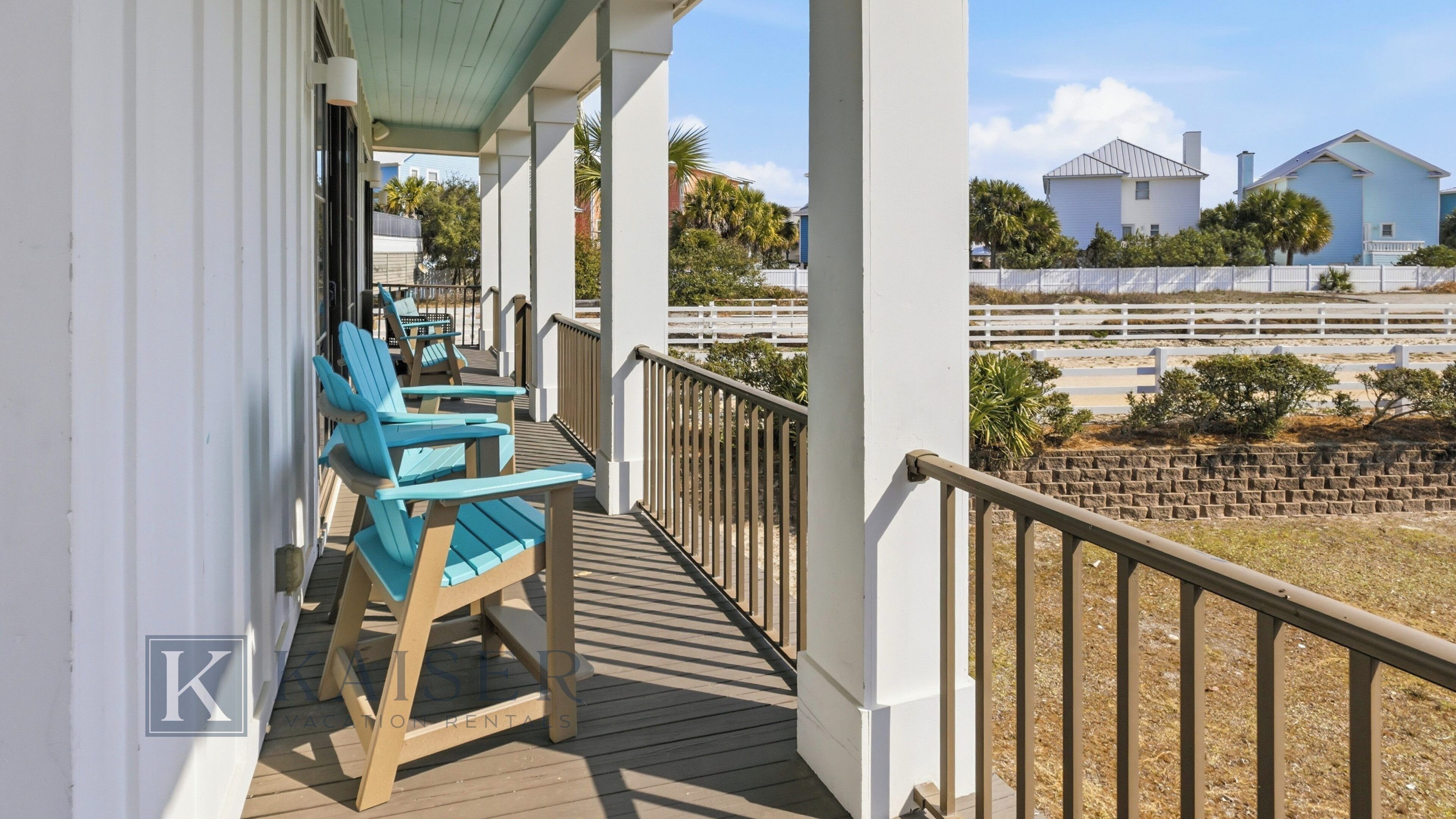 A wooden deck with two blue chairs, a white railing, and a view of houses and a fence in the background.