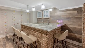 A kitchen with a central island, bar stools, pendant lighting, and a neon sign. - Ping-pong | 10-min to NC State | Fire Pit | BBQ (Raleigh)