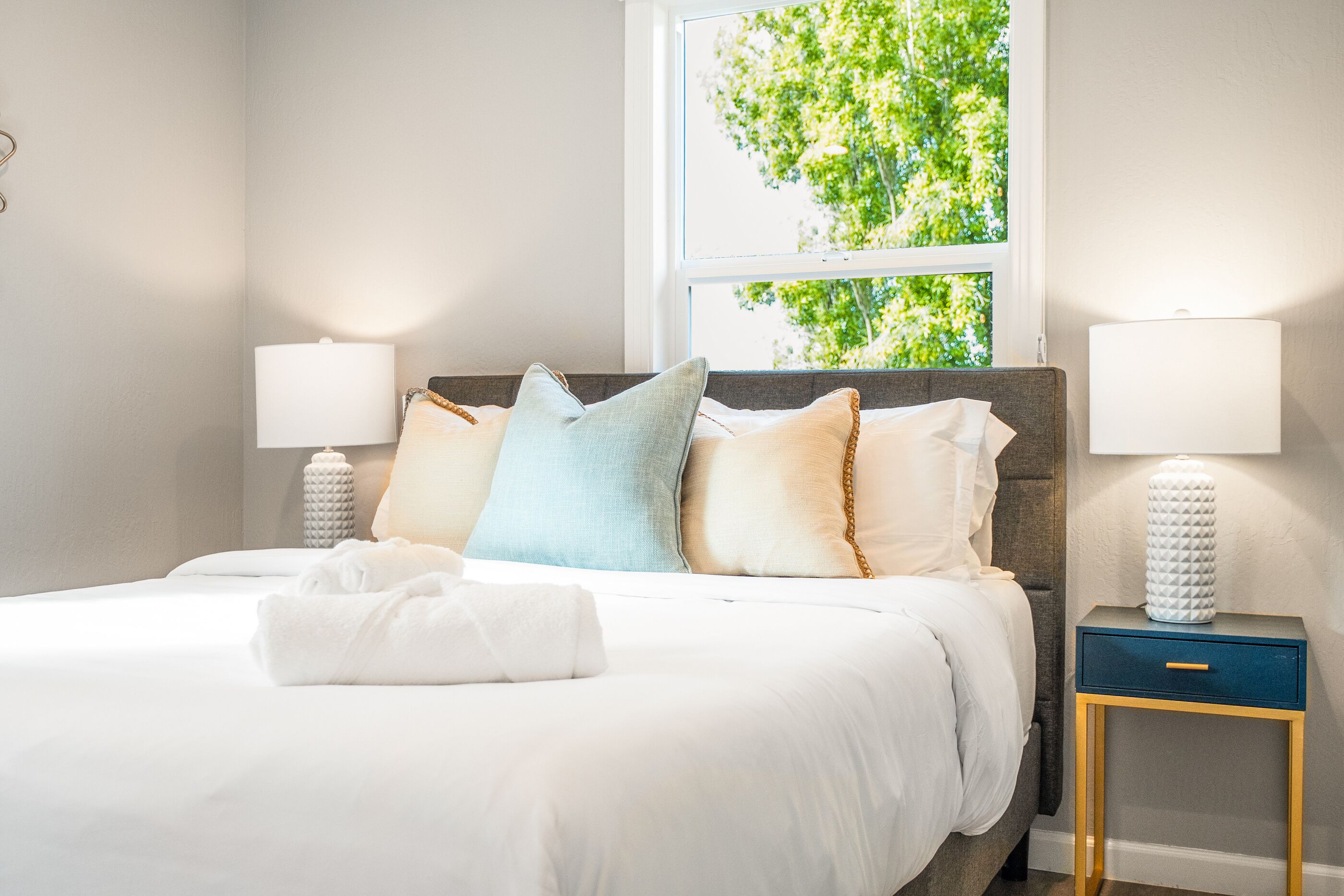Una cama bien hecha con sábanas blancas y varias almohadas, una mesita de noche con una lámpara, y una ventana con vistas a árboles.