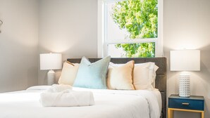 Una cama bien hecha con sábanas blancas y varias almohadas, una mesita de noche con una lámpara, y una ventana con vistas a árboles.