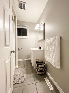 Une salle de bain avec un lavabo blanc, un miroir et un porte-serviettes. Il y a une fenêtre au-dessus du lavabo et une porte sur la gauche.