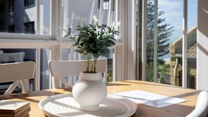 A white vase with flowers on a wooden table with a white placemat, near a window with a view of buildings and trees. - Seaside apartment, 100m to beach (Narrabeen)