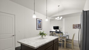 Cozinha moderna com bancada de mármore, armários de madeira, área de jantar com mesa e cadeiras, e televisão embutida.