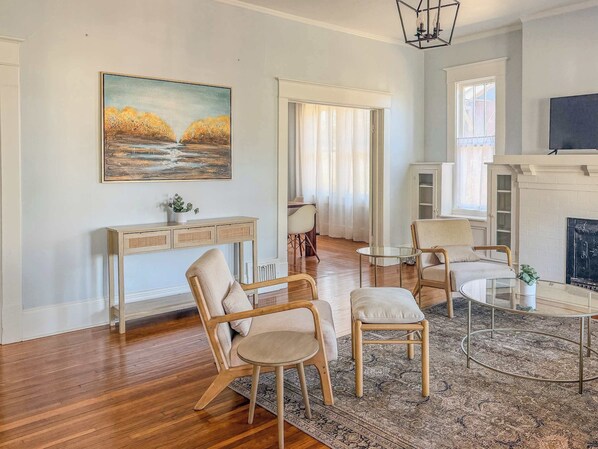 A living room with a wooden floor, a sofa, two armchairs, a glass coffee table, a console table with a plant, a painting, a fireplace, and a TV.