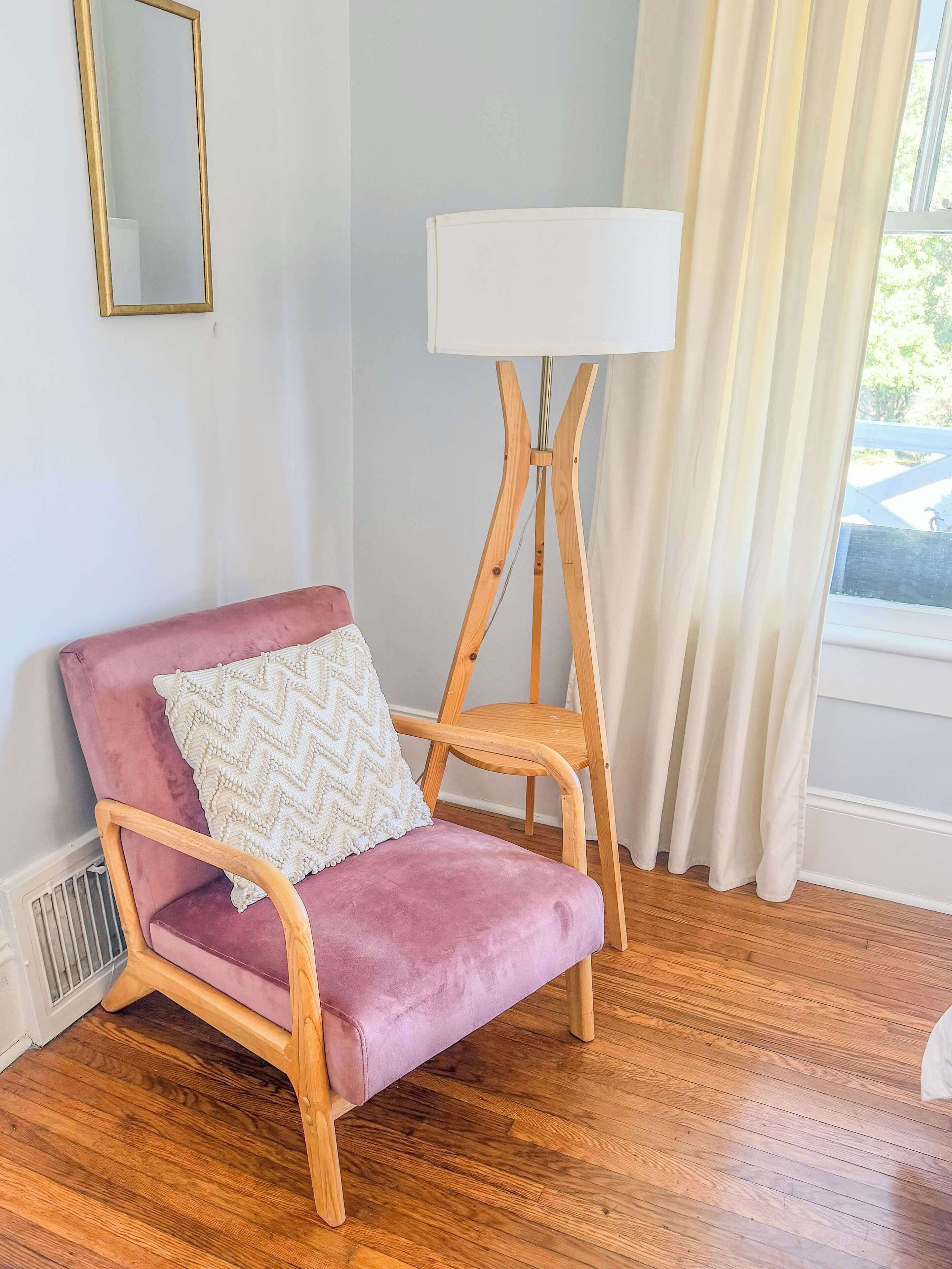 Une pièce avec une chaise rose, une lampe blanche et une fenêtre avec des rideaux.