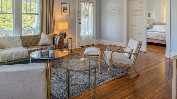 Een ruime woonkamer met een bruine leren bank, een glazen salontafel en een kamerbreed tapijt. Er is een deur naar een slaapkamer met een bed en nachtkastje.
