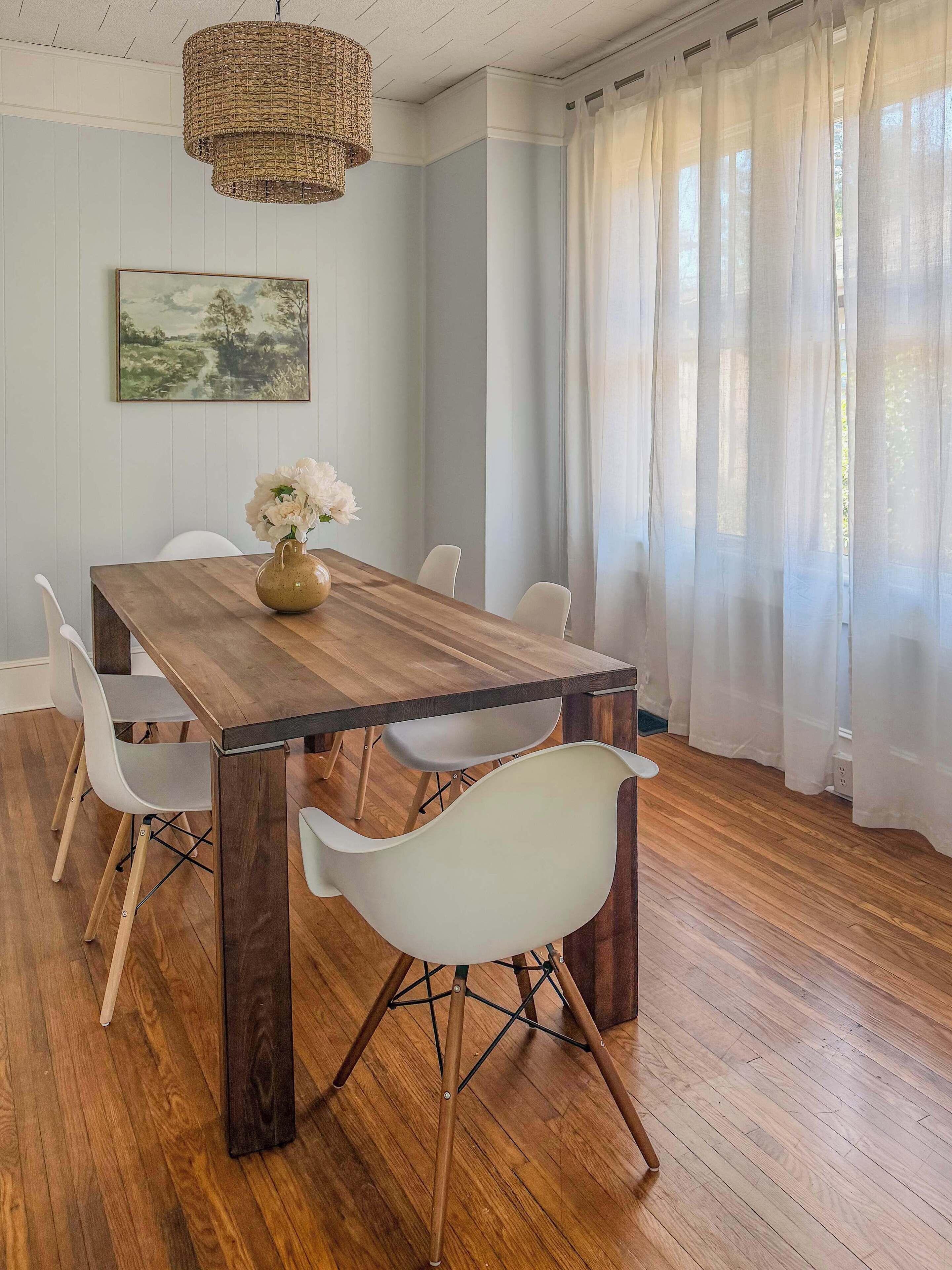 Een eethoek met een houten tafel, witte stoelen, een bloevas en een hanglamp.