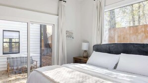 Un dormitorio con una cama grande, una ventana con cortinas, una mesita de noche con lámpara y vistas al exterior.