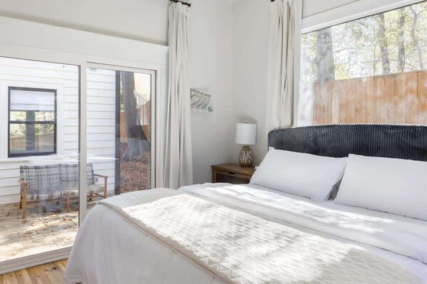 A bedroom with a large bed, a window with curtains, a nightstand with a lamp, and a view of the outdoors. - Designer Garden Home Downtown (Durham)
