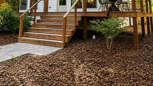 A wooden deck with a staircase leading to a covered porch area, surrounded by trees and a mulched ground. - Designer Garden Home Downtown (Durham)