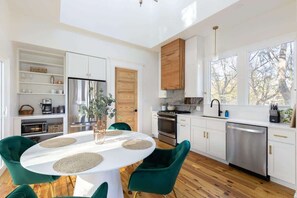 A modern kitchen with white cabinetry, a wooden door, and a dining area with a white table and green chairs. - Designer Garden Home Downtown (Durham)