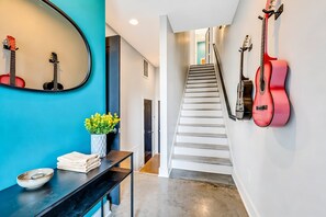 A staircase leading to a second floor, a wall-mounted guitar rack, a mirror reflecting guitars, a vase with yellow flowers, and a black console table with towels and a bowl.