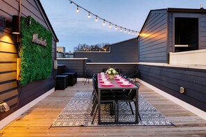 A rooftop patio with a long table set for dining, surrounded by chairs and a patterned rug.