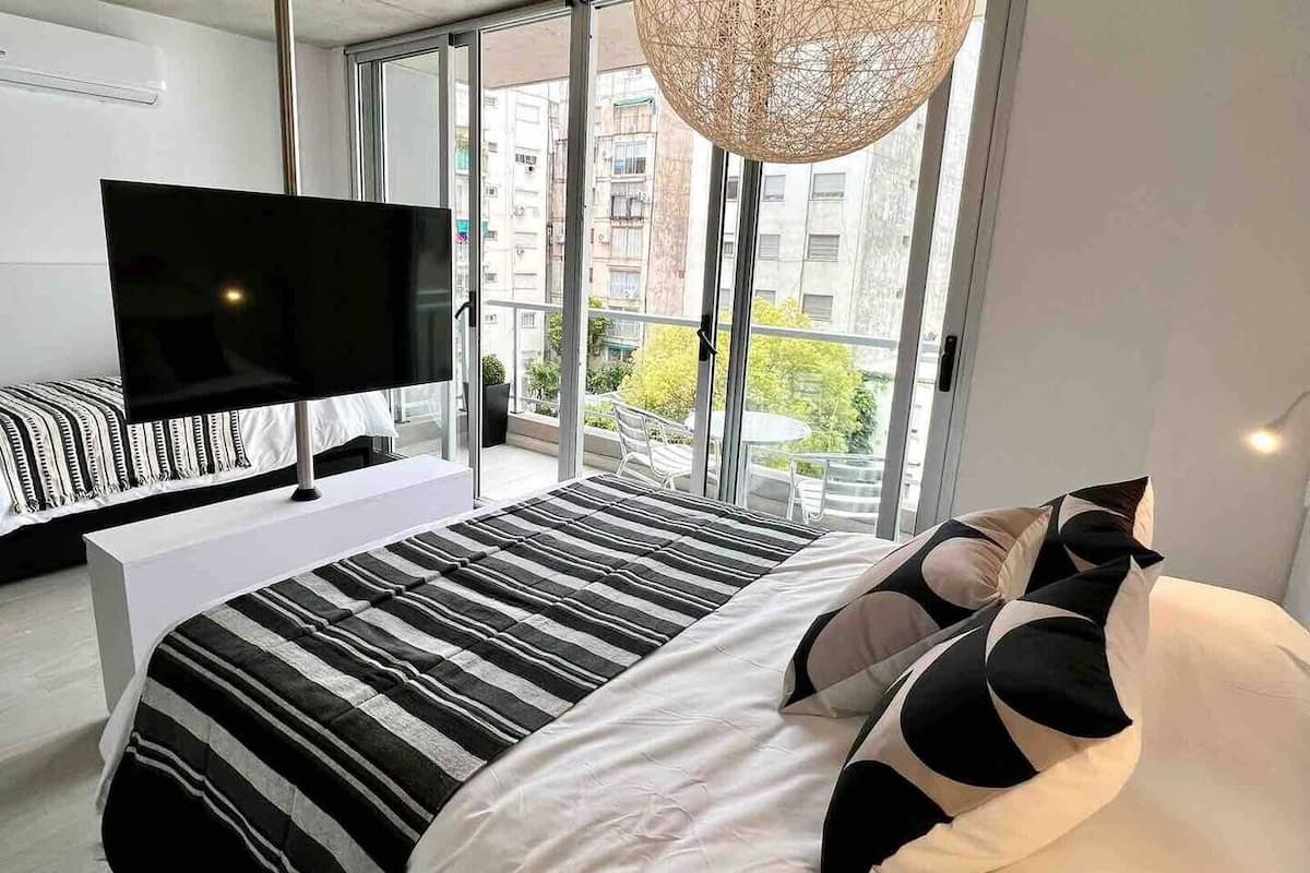 A modern bedroom with a large bed, a flat-screen TV, and a view of the city through large windows.