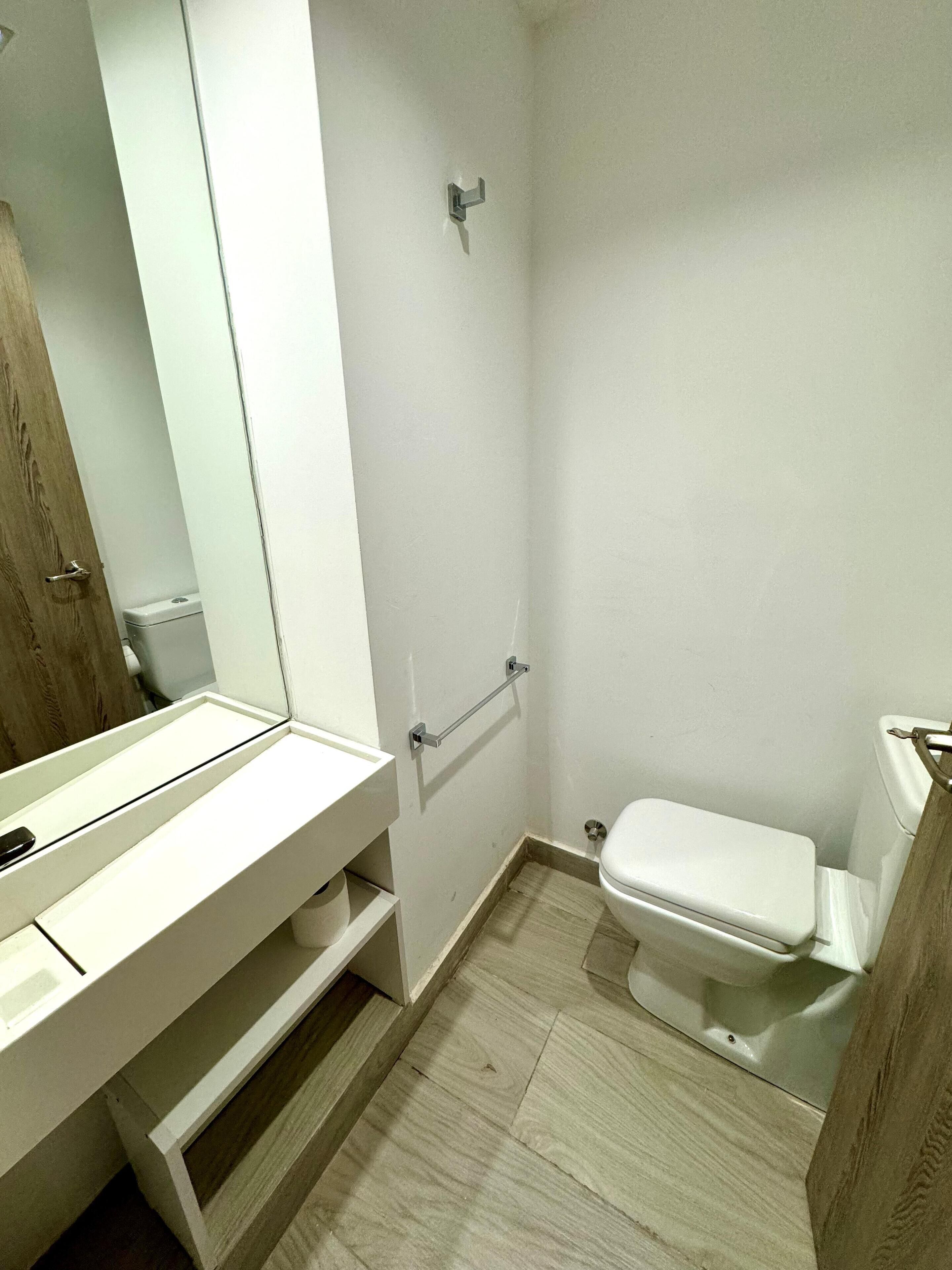 Ein kleines, sauberes Badezimmer mit Toilette, einer Holztür und einem weißen Waschbecken mit Spiegel.