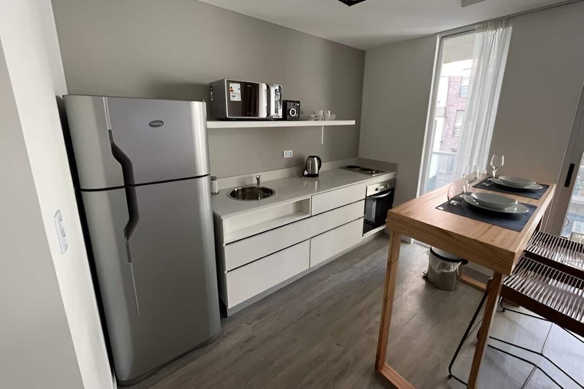 A modern kitchen with a built-in refrigerator, a microwave, and a kettle on a white countertop. There's a wooden table with a grill on the right.