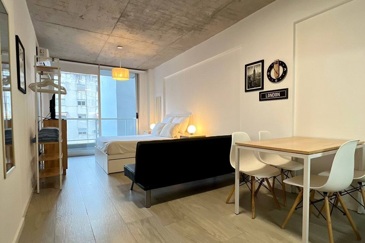 A modern living room with a sofa, dining table, and chairs. There are clocks on the wall and a view of the outside.