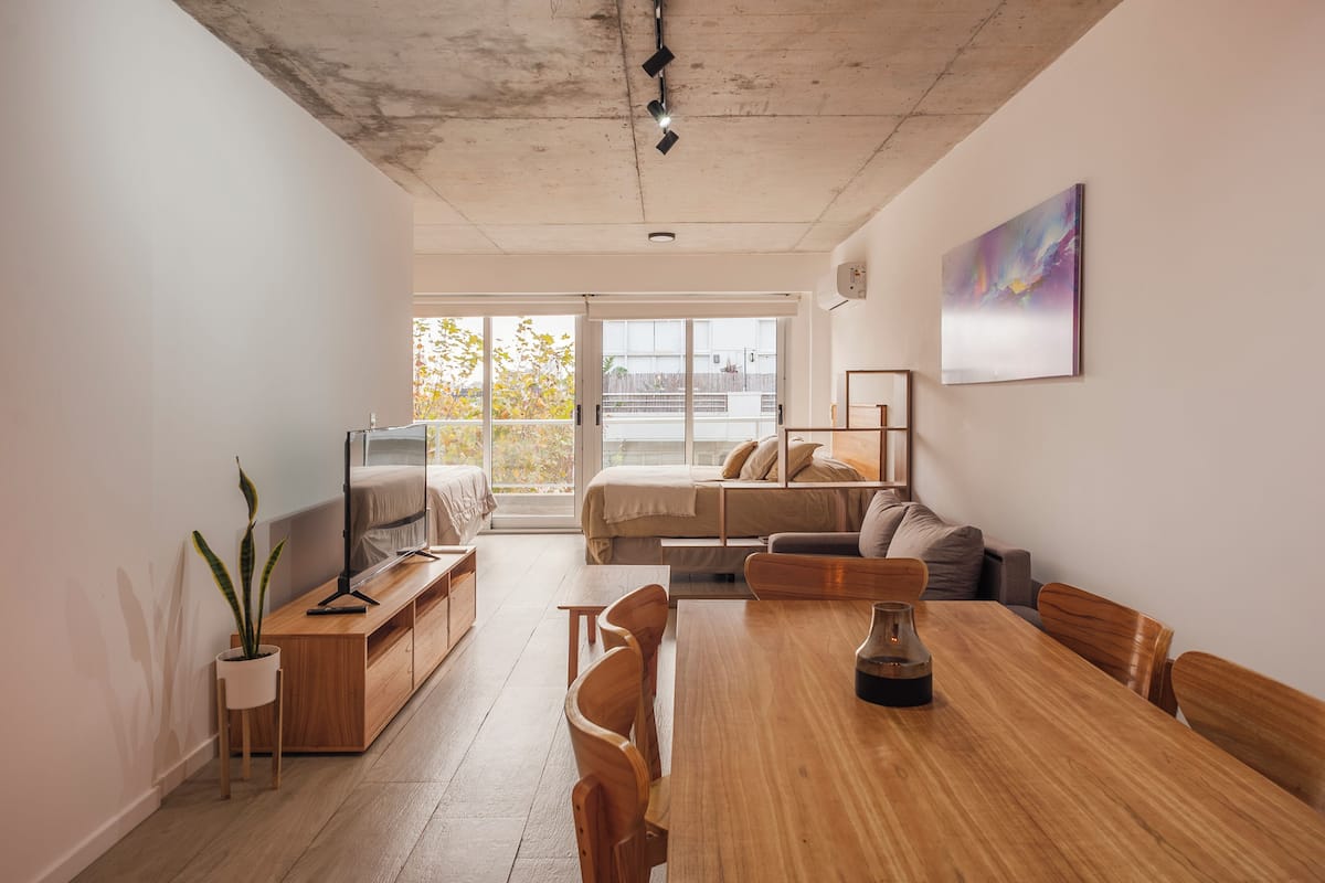 A modern living room with a wooden dining table, sofas, a TV, and a balcony with a view.
