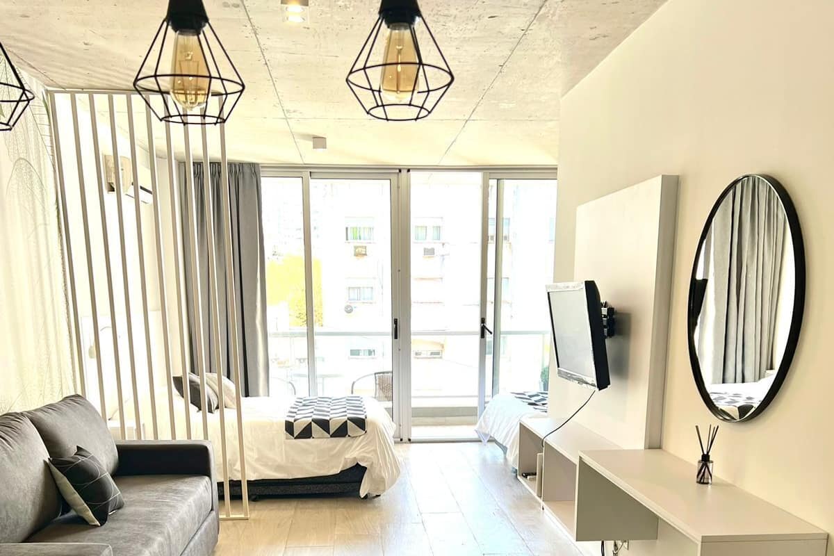 A modern hotel room with a sofa, a dining table with chairs, a TV, a mirror, and a large window with a view of the city.