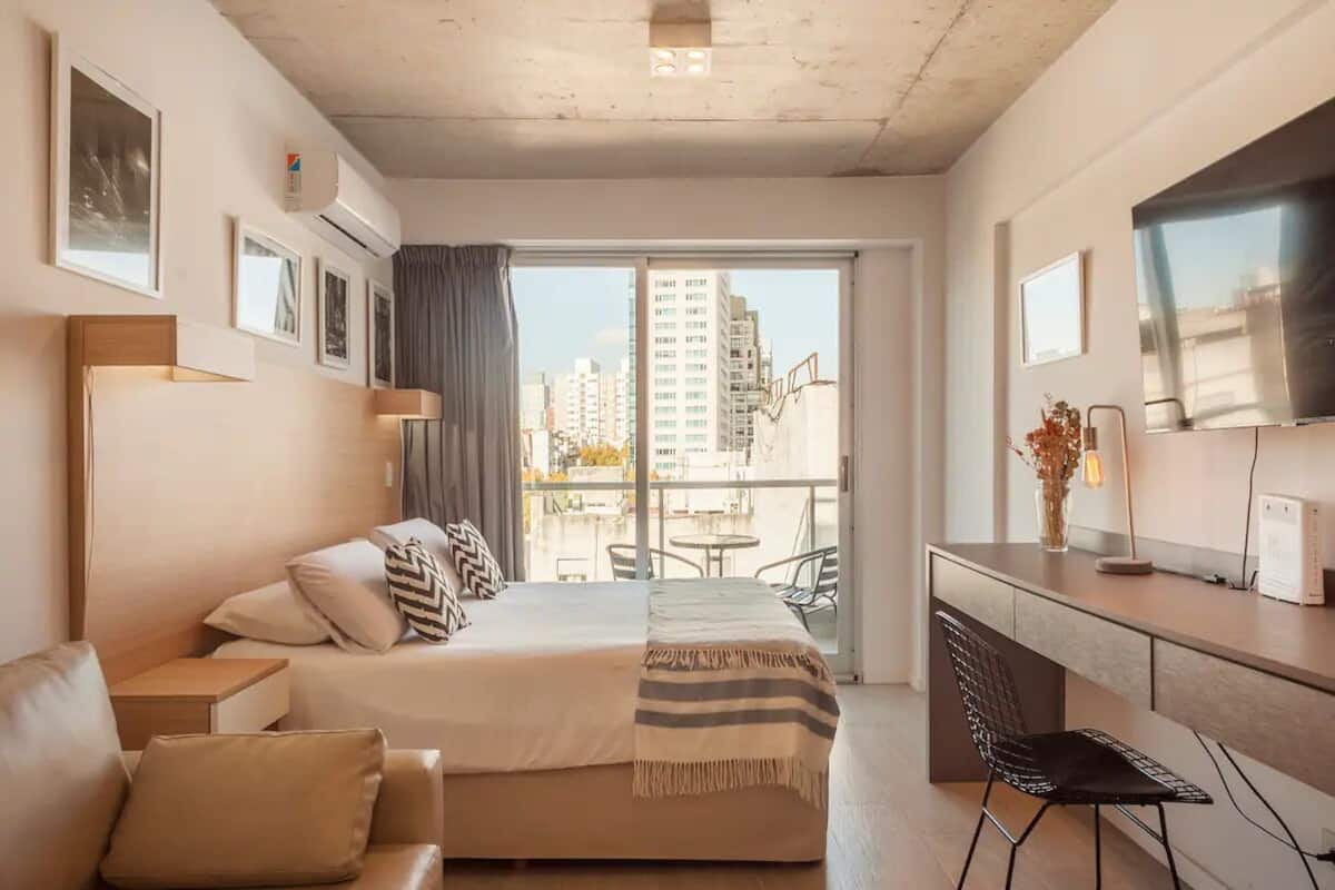A modern hotel room with a bed, a desk, a chair, and a balcony with a city view.