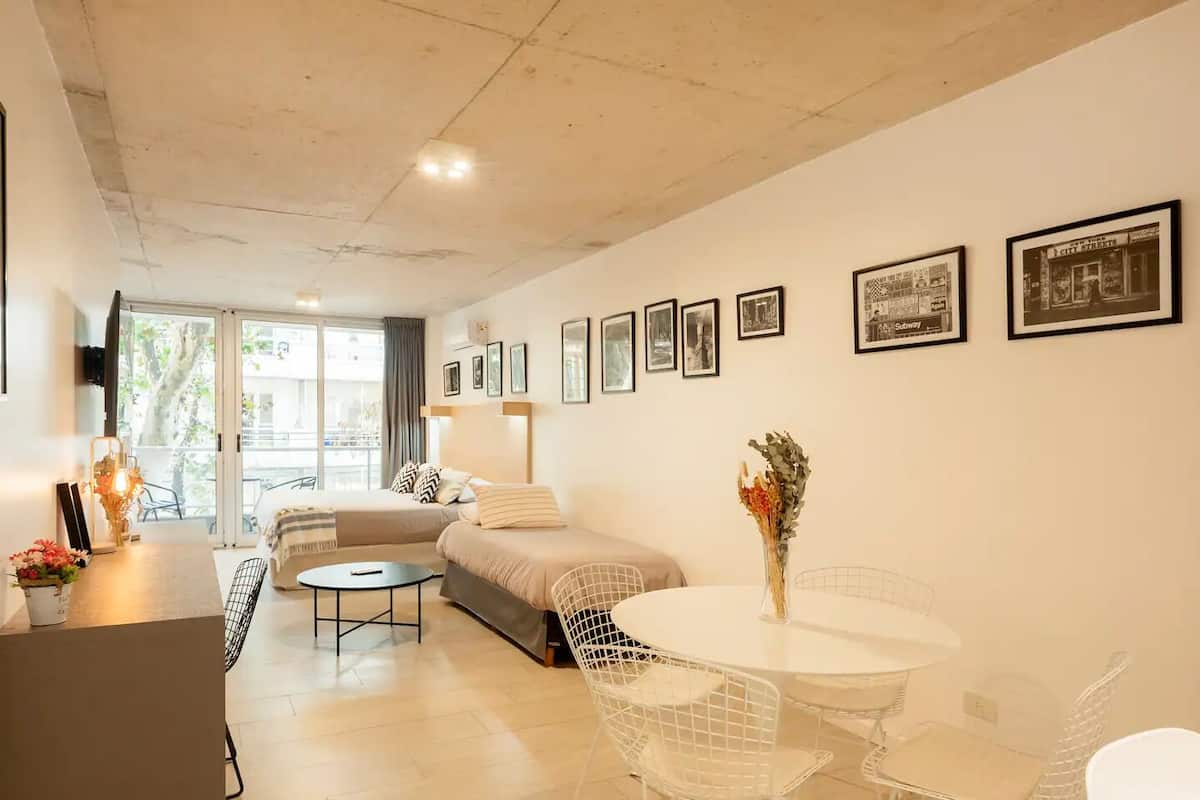 A modern hotel room with a sofa bed, a dining table with chairs, a small kitchen area, and a balcony with a view.