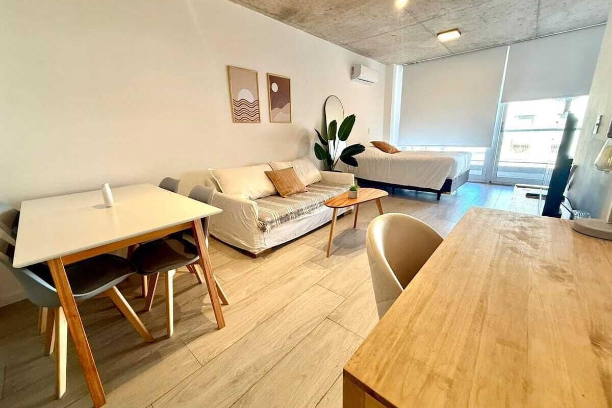 A modern living room with a sofa, a dining table, and a coffee table. There is a large window providing natural light and a view of the outside.
