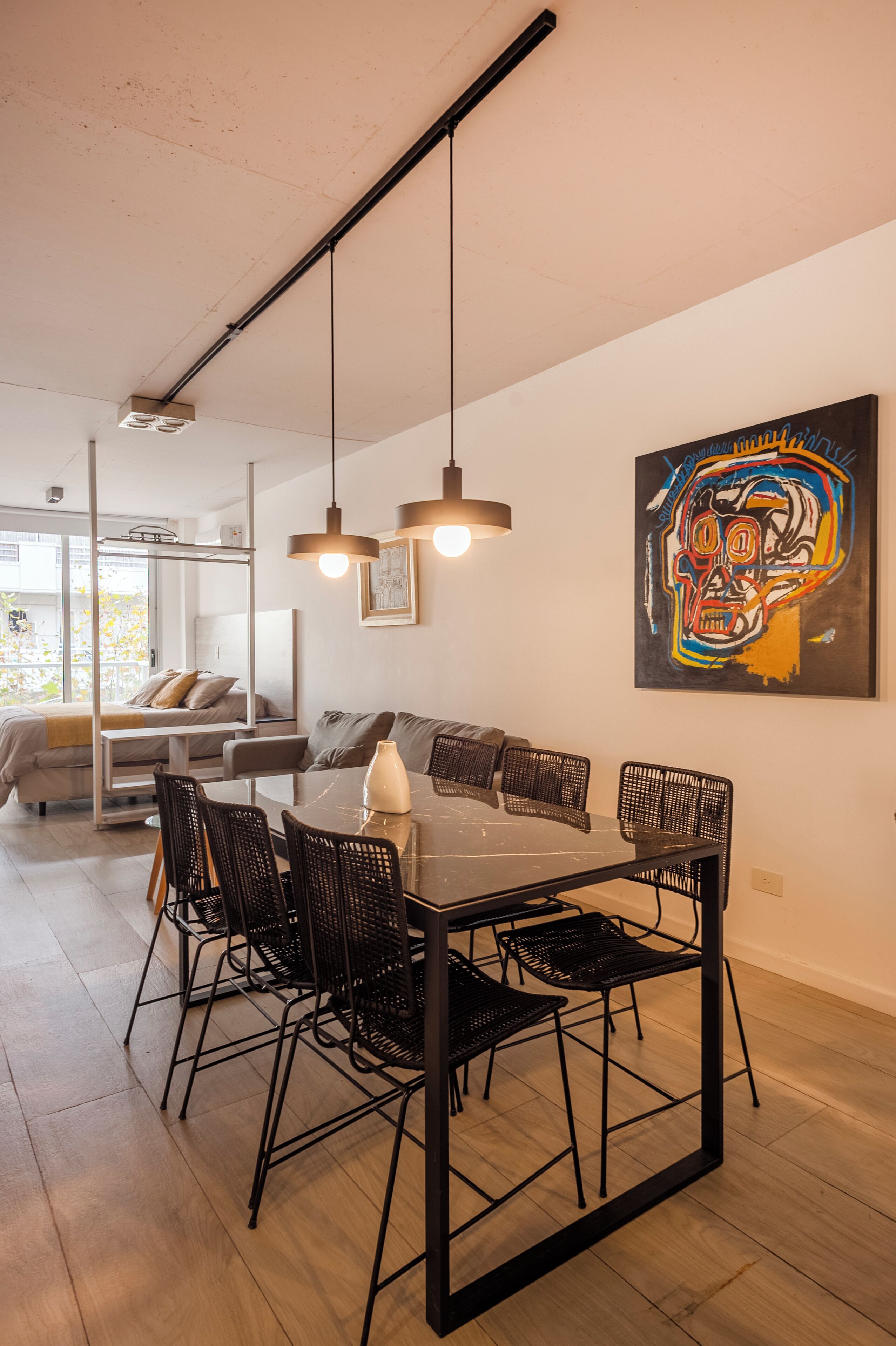 A modern dining area with a black metal table and chairs, a hanging light fixture, and a colorful abstract painting on the wall.