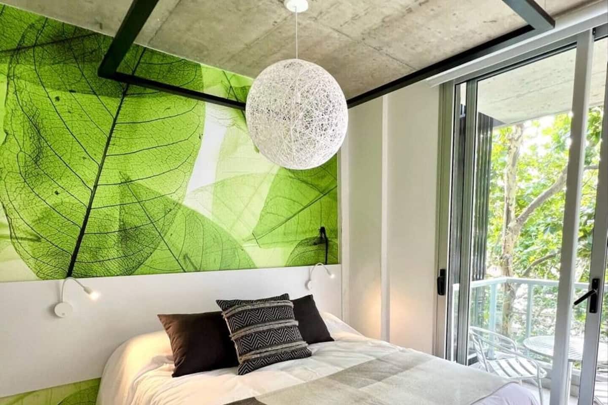 A modern bedroom with a large bed, a green leaf-patterned wall, a white ceiling, and a balcony with a view of trees.
