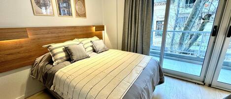 Une chambre à coucher avec un grand lit, une tête de lit en bois et une vue sur le paysage urbain, visible à travers une porte-fenêtre coulissante.