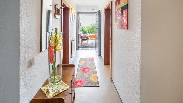 Un pasillo con un banco de madera, un jarrón con flores, un espejo y una puerta que da a un espacio exterior.