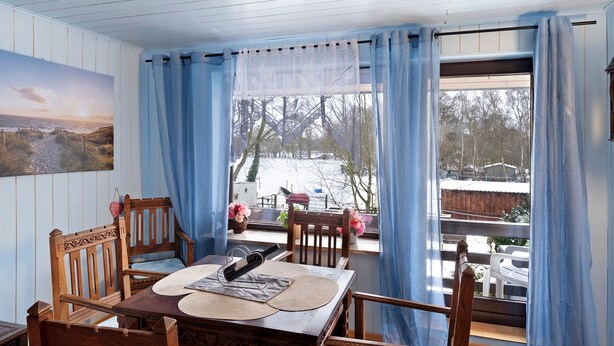 A dining area with a wooden table, chairs, and a view of the outdoors through a window with blue curtains. - Holiday Apartment 'Mole Valley Ranch 2' with Balcony and Wi-Fi (Elmlohe)