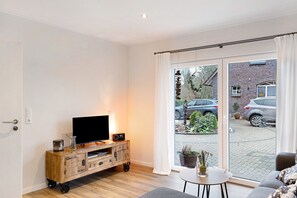 Un salon moderne doté d’un téléviseur, d’un meuble de rangement en bois et offrant une vue sur la rue grâce à de grandes portes vitrées.