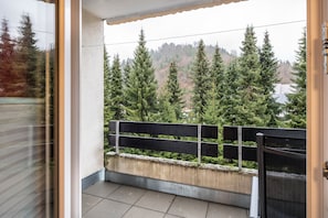 Un balcon doté d’une rambarde en métal, d’une corniche en béton et offrant une vue sur des arbres à feuilles persistantes et un paysage vallonné.