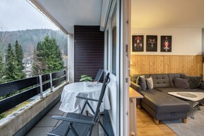 Um espaço de estar moderno com varanda, sofá, mesa de jantar e vista para árvores e colinas.