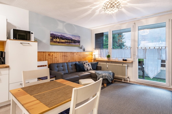 A modern kitchen with a microwave, a dining table with chairs, a sofa, and a large window with curtains.