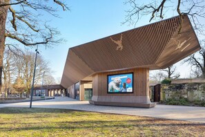 A modern building with a unique sloped roof design, a large digital billboard advertising a zoo, and a stone wall to the right.