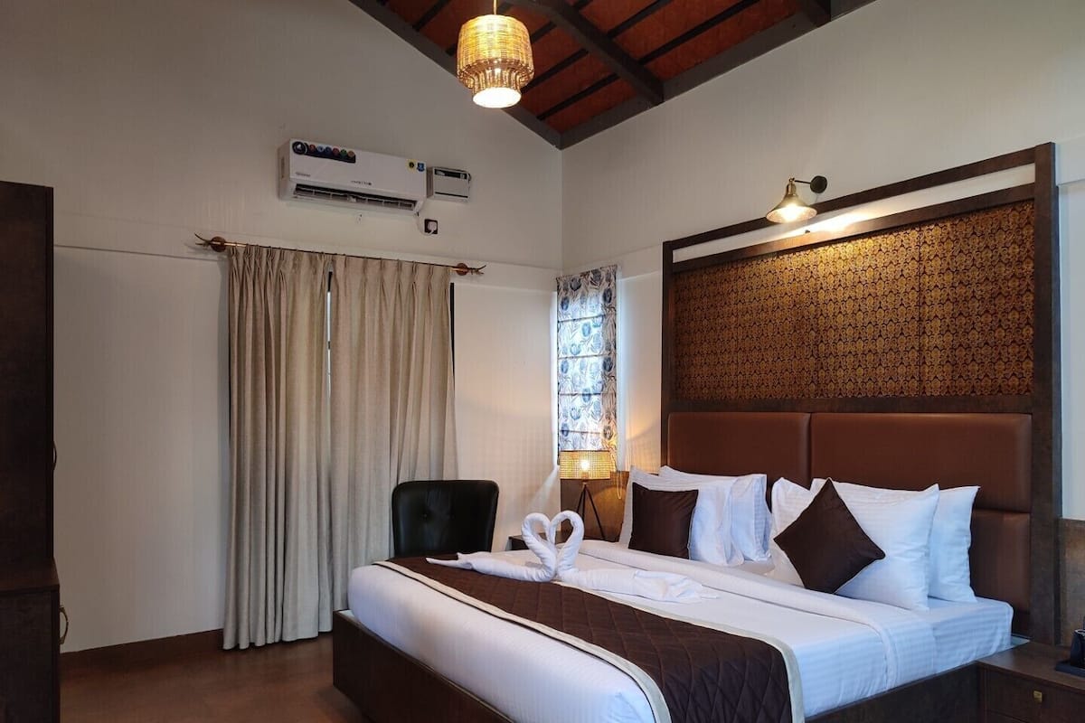 Quarto de hotel com uma cama grande, cabeceira de madeira, ar condicionado fixado na parede, janela com cortinas e luminária pendente.