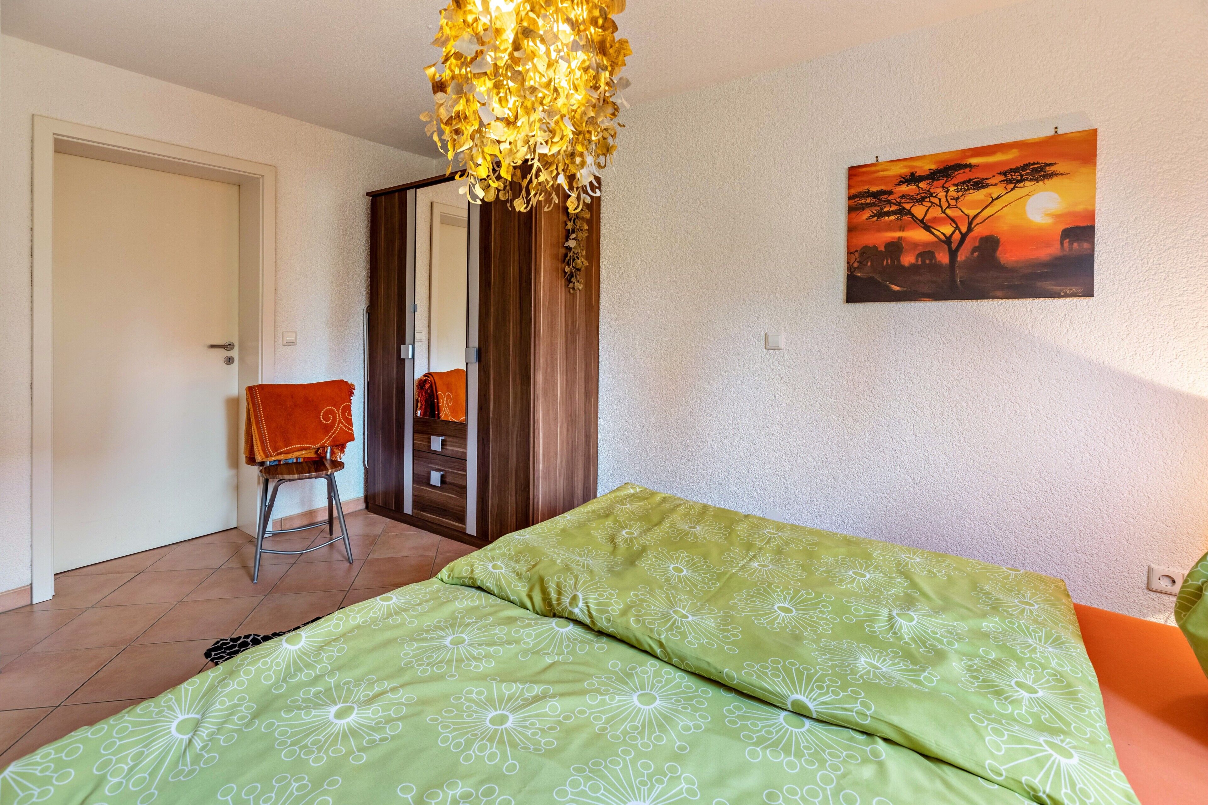 A bedroom with a bed, a chair, a wardrobe, a painting, and a hanging light fixture.