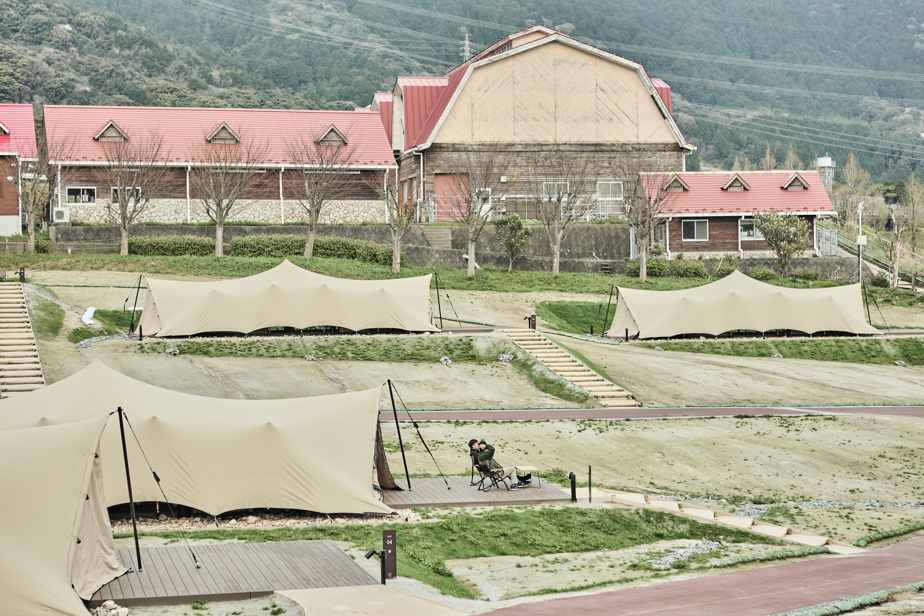 A campsite with tents, a wooden deck, and a large building with a red roof.