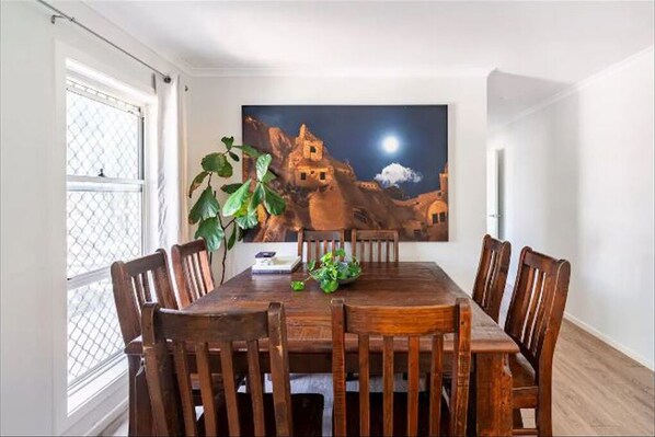 A dining area with a wooden table and chairs, a large painting of a mountain scene, and a potted plant. - North Creek Family Escape (Ballina)