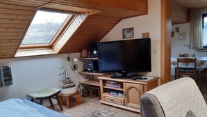 Una camera da letto mansardata accogliente, con soffitto spiovente, un'ampia finestra, un letto, una scrivania con un computer e un televisore.