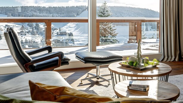 Una habitación moderna con un ventanal grande, un sofá, un sillón y una mesita con una planta.