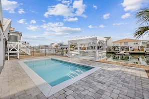 Une piscine entourée d’une terrasse en briques, agrémentée de pergolas blanches, et surplombée d’un ciel d’un bleu limpide.