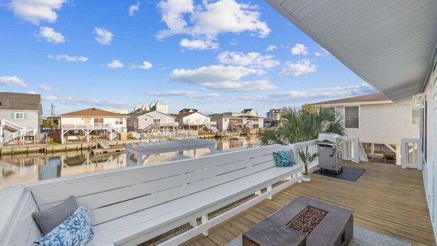 A waterfront deck with a wooden bench, a grill, and a view of houses across the water. - 5BR Waterfront w/ Heated Pool | Fire Pit | Grill (North Myrtle Beach)