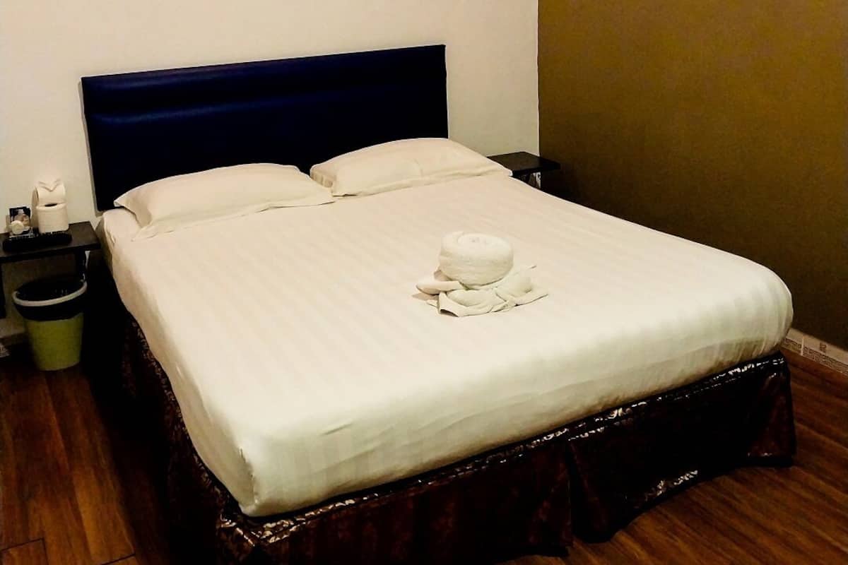 Una cama bien hecha con una colcha blanca y una toalla decorativa, un cabecero oscuro, una mesita de noche con una lámpara, un cesto de basura y un aire acondicionado instalado en la pared.